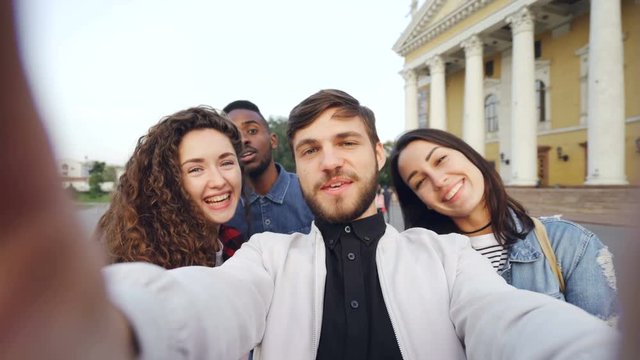 Point of view shot of multiracial group of tourists taking selfie in city center holding camera and posing together with hand gestures expressing positive emotions.