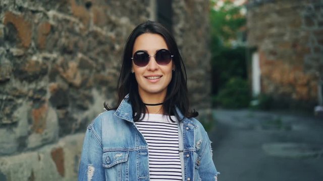 Slow motion portrait of beautiful young woman in sunglasses and denim jacket standing outdoors with brick walls in background, smiling and looking at camera.