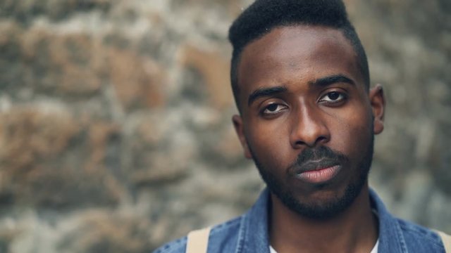Close-up slow motion portrait of emotional African American man looking at camera with straight face then smiling happily reacting to good news. People and emotions concept.