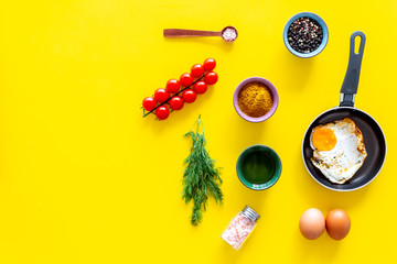 Recipe of fried eggs with vegetables. Ready eggs in a frying pan near cherry tomatoes, greenery, spices, raw eggs on yellow background top view copy space