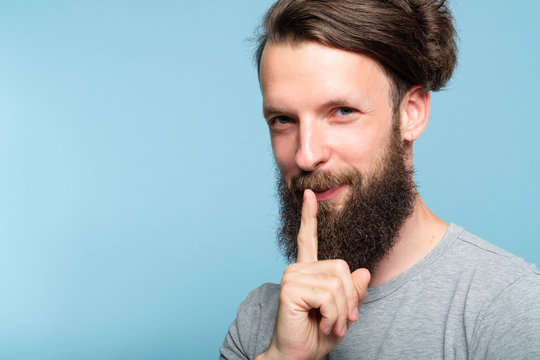 smiling smug man keeps a secret. confidential information and business tips sharing concept. portrait of a bearded hipster guy on blue background.