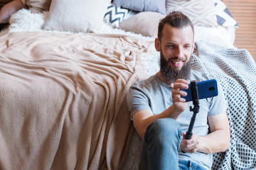 bearded hipster man taking a selfie using phone camera on a stick. idle leisure and relaxed carefree lifestyle. guy sitting at home near the bed. modern social trends concept.