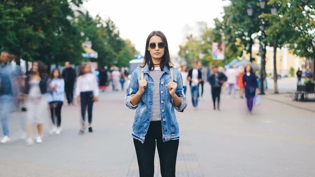 Zoom out time-lapse of confident young lady in sunglasses looking at camera standing in busy pedestrian street in flow of people. Youth and society concept.