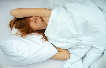 Portrait einer attraktiven, zufriedenen, jungen, sexy, rothaarigen Frau, die entspannt im Bett liegt, genießt und kuschelt mit frischer, weicher, weißer Bettwäsche und Kissen im Schlafzimmer.