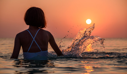 Young girl playing with water splashes under the first rays of the rising sun at beach in Greece -...