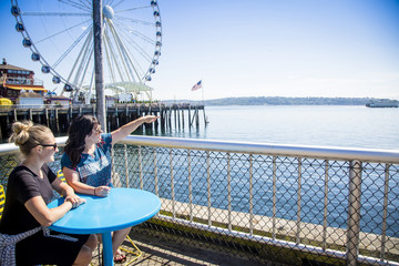 Two women looking out at the Seattle harbor on a sunny day. Sitting at a table on the waterfront near the Ferris Wheel and pier - obrazy, fototapety, plakaty
