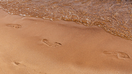 Footprints of bare feet in the sand along a river bank with an oncoming wave