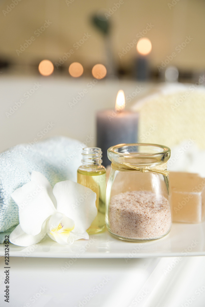 Wall mural White ceramic tray with home spa supplies in home bathroom for relaxing rituals. Candlelight, salt soap bar, bath salt in jar, massage, bath oil in bottle, blue rolled towel, natural sponge. 