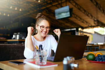 Portrait of happy young business woman celebrating success with arms up in front of laptop. Mixed...