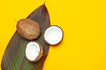 Ripe coconut and tropical leaves on yellow colored background, minimal flat lay style top view. Pop art design, creative summer and food concept. Tropical fruit whole and half abstract background