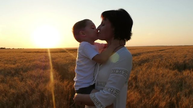 A happy mother is holding her child in her arms in a field of wheat against the background of a sunset. A happy child kisses the mother's face, mother kisses the child