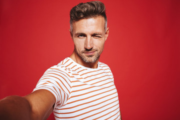 Fototapeta na wymiar Caucasian guy 30s in striped t-shirt smiling while taking selfie photo, isolated over red background