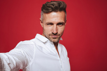Obraz premium Attractive bearded man 30s in white shirt taking selfie photo, isolated over red background