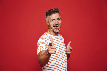 Smiling adult man in striped t-shirt gesturing index finger on camera meaning hey you, isolated...