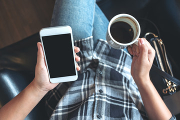 Top view mockup image of woman's hands holding white mobile phone with blank black desktop screen while drinking coffee in cafe