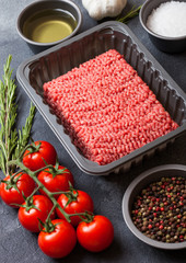 Tray with raw minced homemade beef meat with spices and herbs. Top view. On top of kitchen table background. With pepper salt and tomatoes.