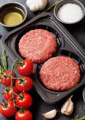 Plastic tray with raw minced homemade beef burgers with spices and herbs. Top view. On top of wooden kitchen table background with tomatoes salt and pepper.