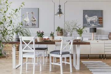 White chairs at table with flowers in rustic dining room interior with lamp and posters. Real photo