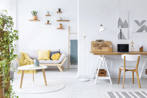 Posters above desk with laptop in white living room interior with couch and wooden table. Real photo