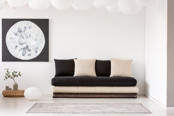 Pillows on black sofa near carpet in white living room interior with plant and moon poster. Real...