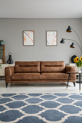 Blue patterned carpet and leather sofa in grey living room interior with posters and lamp. Real photo