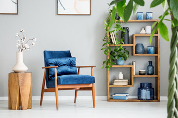 Blue armchair next to wooden table with white flowers in grey flat interior with plants. Real photo
