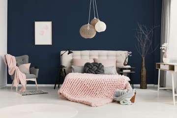 Real photo of knot cushion and pastel pink coverlet placed on double bed in elegant hotel room interior with simple poster on dark blue wall, armchair in the corner and twigs in tall vase