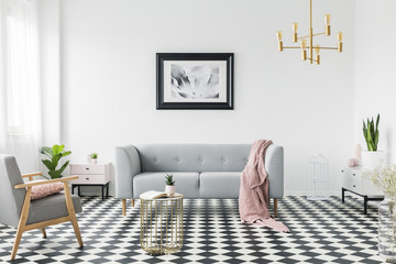 Grey sofa with pink blanket under poster in white flat interior with armchair and gold table. Real photo