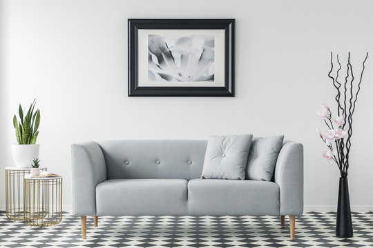 Poster above grey couch with pillows in minimal living room interior with plant on gold table. Real photo