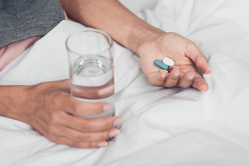 cropped shot of sick woman holding pills and glass of water while lying in bed