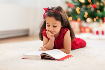 christmas, holidays and childhood concept - happy little girl lying on floor and reading book at...