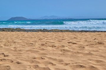 Fototapeta na wymiar Landscape of golden sand and stones on the shore with the sea and waves and mountains in the background on a clear day in Corralejo, Fuerteventura, Canary Islands, Spain