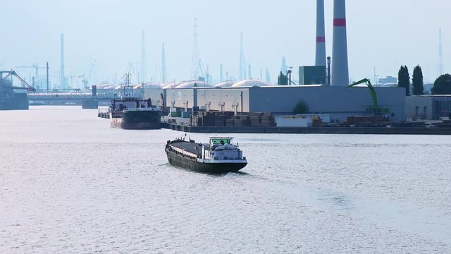 Loaded barge passing by Antwerp international seaport at  Scheldt river. Silhouettes of cranes, rotating wind turbines and stacked cargo containers near warehouse buildings on quayside. Belgium