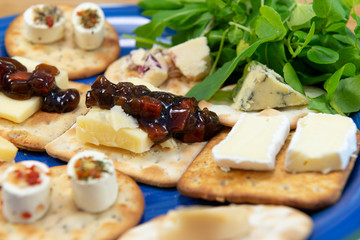 Crackers topped with a selection of cheeses, with sweet pickle and salad leaves. Close up view with focus on pickles. On a blue plate.