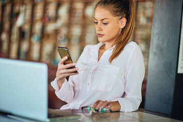 Businesswoman sitting in restaurant and using smart phone. On table eyeglasses and laptop.