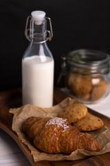Croissant with coconut chips on kraft paper, next to a transparent bottle of milk and a transparent jar of cookies on black background