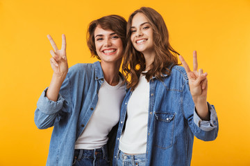Women friends posing isolated over yellow background showing peace gesture.