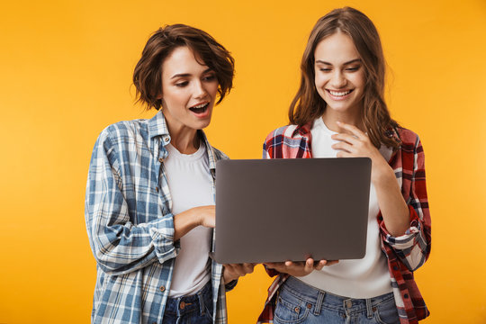 Women friends posing isolated over yellow background using laptop computer.