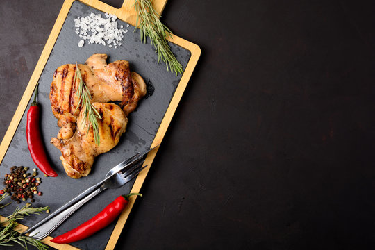 Grilled chicken fillets on slate plate with rosemary, pepper and spices on dark wooden background. Top view. Flat lay. Copy space