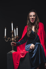 dreadful woman in red cloak holding vintage candelabrum and showing vampire teeth isolated on black