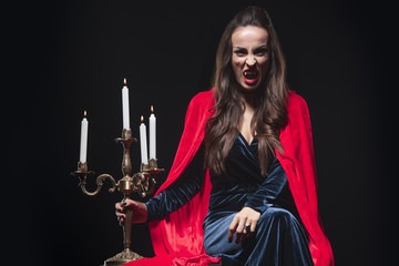 woman in red cloak holding vintage candelabrum and showing vampire fangs isolated on black