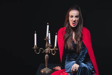sexy woman in vampire costume sitting near vintage candelabrum isolated on black