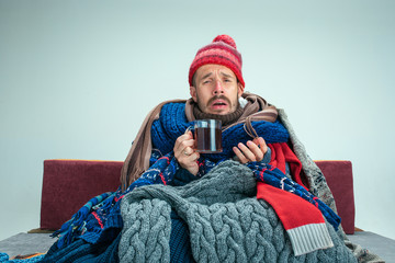 Bearded sick man with flue sitting on sofa at home or studio with cup of tea covered with knitted...