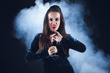 grim vampire holding wineglass with blood on dark background with smoke