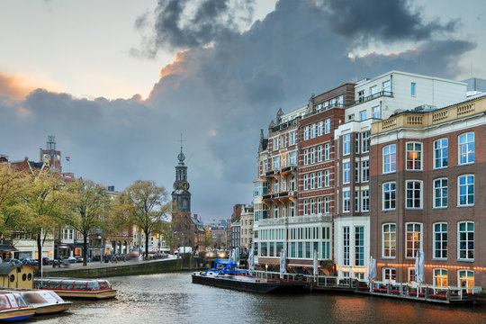 Beautiful cityscape view towards the Mint tower (Munttoren) with tourist canal boats at the Amstel river in Amsterdam, the Netherlands, with ominous clouds around sunset