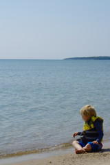 Fototapeta na wymiar Cute Young Blonde School Aged Boy Sitting on Sand Beach Reaching Out Toward Water