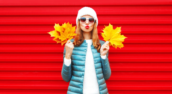 Fashion autumn portrait woman holds yellow maple leaves, blows red lips, sends an air kiss on background in city