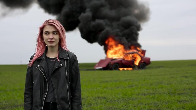 Young beautiful emo-girl with pink hair and in a leather jacket stands on the background of an exploding car, a hipster and a car explosion