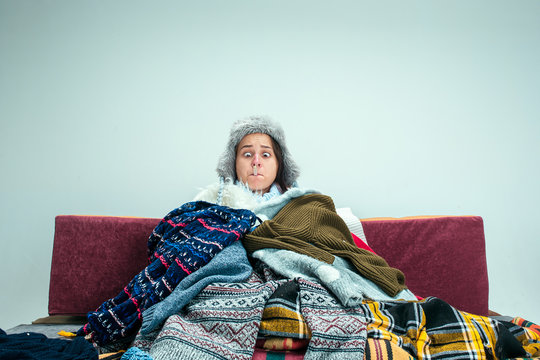 The young sick woman with flue sitting on sofa at home or studio covered with knitted warm clothes. Illness, influenza, pain concept. Relaxation at Home. Healthcare Concepts.