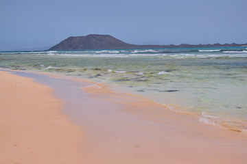 Fototapeta na wymiar Paradisiacal landscape with sand and beach with waves in the foreground and mountain background on a sunny day in Corralejo, Fuerteventura, Canary Islands, Spain